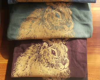 May include: A stack of four folded t-shirts in blue, green, maroon, and black. Each shirt features a screen-printed graphic of a rabbit head in a mustard yellow ink, showcasing detailed line work and texture on the animal's fur and ears.