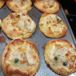 May include: A close-up of a muffin tin filled with golden brown chicken pot pie bites. The bites are topped with a creamy sauce and vegetables.