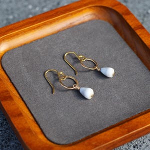 May include: A pair of gold-colored earrings with a teardrop-shaped, pale blue bead. The earrings are displayed on a gray surface inside a wooden tray with a dark brown finish.