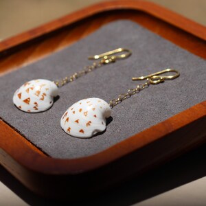 May include: A pair of drop earrings featuring white seashells with small gold triangular patterns. Each shell is attached to a thin gold chain and a gold hook ear wire. The earrings are displayed on a gray fabric insert within a wooden tray.