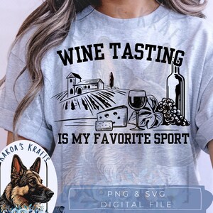 May include: A light gray short-sleeve t-shirt featuring a black graphic print of a vineyard, a wine glass, a bottle, and cheese. The text reads WINE TASTING IS MY FAVORITE SPORT. A small circular logo shows a German Shepherd dog with blue flowers.