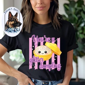 May include: Black t-shirt with a graphic of a smiling dumpling in a yellow bowl, with the text "You're a glitter dumpling" in pink and purple. The shirt also features pink vertical stripes.