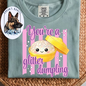 May include: A light blue t-shirt with a graphic of a glitter dumpling in a yellow container, with the text "You're a glitter dumpling" in purple. The shirt also features a logo with a German Shepherd dog.
