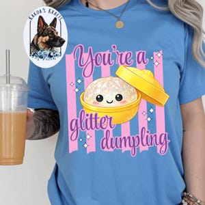 May include: A light blue t-shirt with a graphic of a glitter dumpling in a yellow bowl. The text "You're a glitter dumpling" is written in a cursive font. The shirt also has pink vertical stripes.