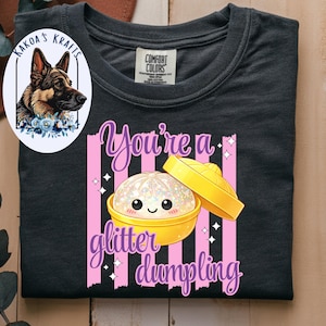 May include: Dark gray t-shirt with a graphic that says "You're a glitter dumpling." The graphic features a cute, glittery dumpling in a yellow bowl with a lid. The shirt also has a logo of a German Shepherd.