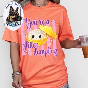 May include: Coral-colored t-shirt with a graphic of a glitter dumpling in a yellow container. The text "You're a glitter dumpling" is written in a whimsical font. A logo with a German Shepherd dog is in the upper left corner.