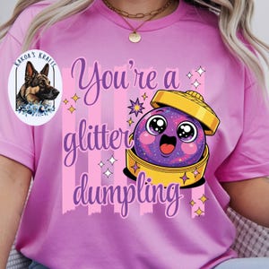 May include: A light pink t-shirt with the text "You're a glitter dumpling" in purple script. The shirt features a cartoon dumpling with a smiling face and a gold-colored lid. The dumpling is purple with glitter.