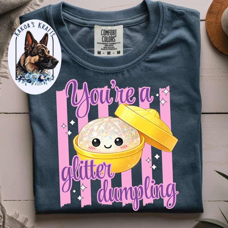 May include: A navy blue Comfort Colors t-shirt featuring a graphic of a glitter dumpling with the text "You're a glitter dumpling". The design includes pink vertical stripes and a cartoon dumpling in a yellow container.
