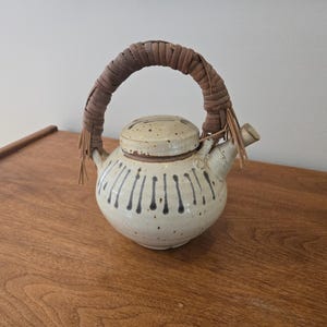 May include: A ceramic teapot with a woven handle and lid. The off-white body is decorated with vertical black lines. The handle is wrapped in brown leather or a similar material. The teapot is on a wooden surface.