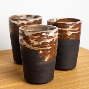 May include: Three handmade ceramic tumblers with a unique design. The cups feature a dark brown base, transitioning to a lighter brown and white speckled pattern near the top. The cups are approximately 5 inches tall.