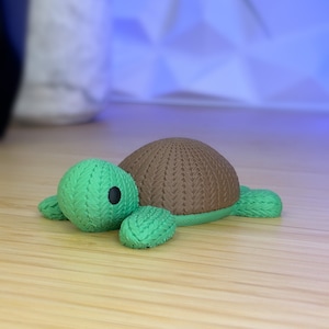 May include: A small, brown and green knitted turtle toy. The turtle has a brown shell and green legs and head.