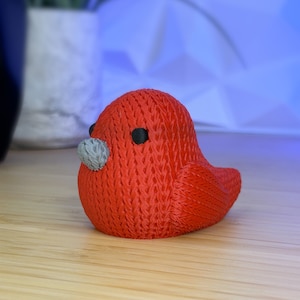 May include: A red 3D printed bird figurine with a gray beak and black eyes. The bird is sitting on a wooden surface.