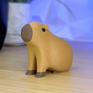 May include: A small, brown, 3D printed capybara figurine with a textured surface. The figurine is sitting on a wooden surface.