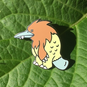 May include: A yellow and brown cartoon platypus enamel pin with a blue beak and feet. The pin is on a green leaf.