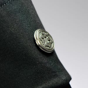 May include: A close-up shot of a silver button with a detailed circular design, fastened to black fabric. The button features a raised, star-like emblem at its centre. The fabric is a dark, woven material.