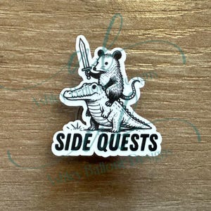 May include: A black and white sticker featuring an opossum riding an alligator and holding a sword. The text "SIDE QUESTS" is below the illustration. The sticker is on a wooden surface.