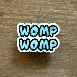 Può includere: Un adesivo bianco con la scritta "WOMP WOMP" in azzurro, bordata di nero. L'adesivo ha una forma arrotondata ed è probabilmente destinato a scopi decorativi.