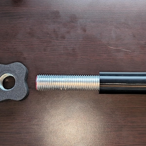 Puede incluir: Una pesa de barra negra y plateada con un mango negro y un peso gris de cuatro lóbulos. La barra tiene un extremo roscado y un acento rojo. El peso está hecho de un material gris oscuro.