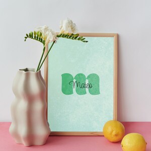 May include: A light green and white framed print with the name "Mateo" in black text. The print is on a light blue background with a subtle texture. The print is sitting on a pink surface next to a white vase and two lemons.