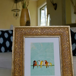 May include: A framed art print featuring a row of colorful birds perched on a thin green line against a light blue background. The frame is ornate, gold-colored, and rectangular. The artwork is displayed in a well-lit interior setting.