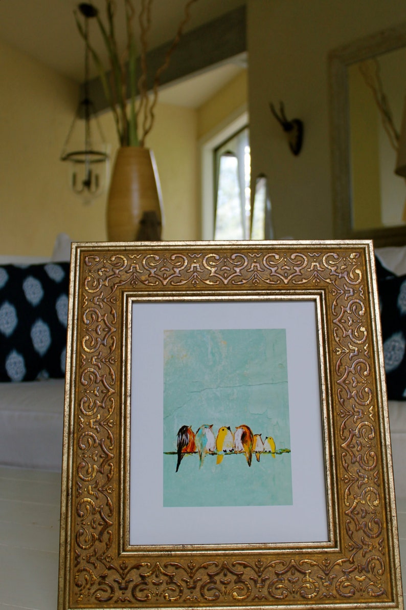 May include: A framed art print featuring a row of colorful birds perched on a thin green line against a light blue background. The frame is ornate, gold-colored, and rectangular. The artwork is displayed in a well-lit interior setting.