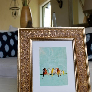 May include: Ornate gold-framed artwork featuring a row of colorful birds perched on a line against a light blue background. The frame has detailed scrollwork. The artwork is displayed on a white surface.
