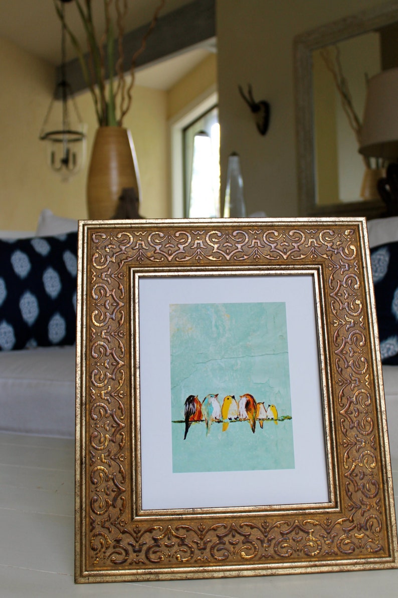 May include: Ornate gold-framed artwork featuring a row of colorful birds perched on a line against a light blue background. The frame has detailed scrollwork. The artwork is displayed on a white surface.