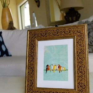 May include: A framed art print featuring a row of colorful birds perched on a branch against a light blue background. The frame is gold with an ornate, repeating pattern. The artwork is displayed on a white surface.