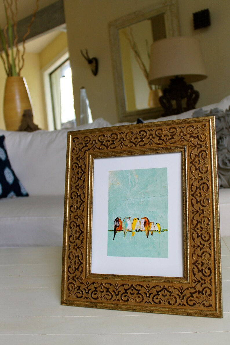 May include: A framed art print featuring a row of colorful birds perched on a branch against a light blue background. The frame is gold with an ornate, repeating pattern. The artwork is displayed on a white surface.