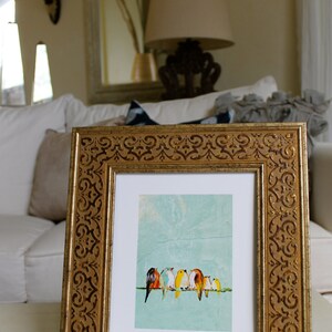 May include: A framed art print featuring a row of colorful birds perched on a wire against a textured, light blue background. The frame is ornate, with a gold finish and decorative detailing. The artwork is displayed on a white surface.