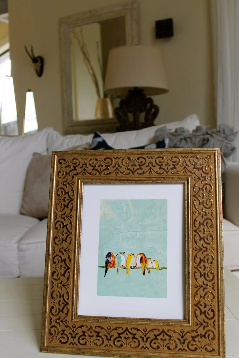 May include: A framed art print featuring a row of colorful birds perched on a wire against a textured, light blue background. The frame is ornate, with a gold finish and decorative detailing. The artwork is displayed on a white surface.