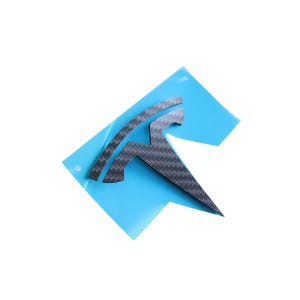 Op de afbeelding: Een zwarte Tesla-logo sticker van carbon fiber op een blauwe ondergrond.