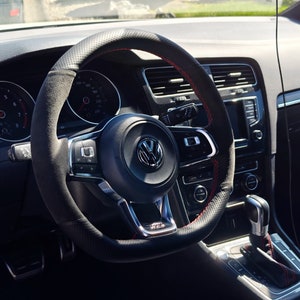 May include: A black leather-wrapped steering wheel with red stitching. The steering wheel has a silver Volkswagen logo in the center and a variety of buttons and controls.