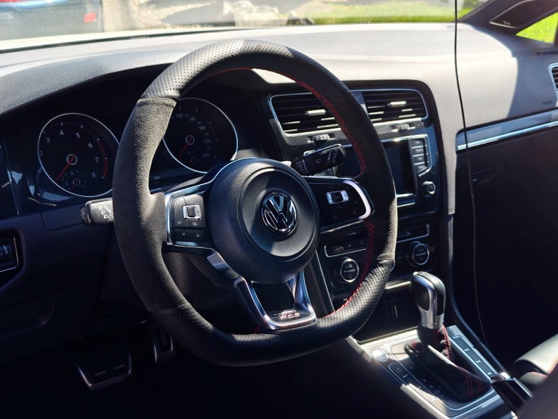 May include: A black leather-wrapped steering wheel with red stitching. The steering wheel has a silver Volkswagen logo in the center and a variety of buttons and controls.