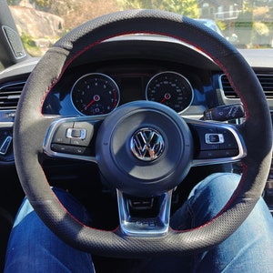 May include: A black leather steering wheel with red stitching and a Volkswagen logo in the center. The steering wheel has controls for the car's audio system and cruise control.