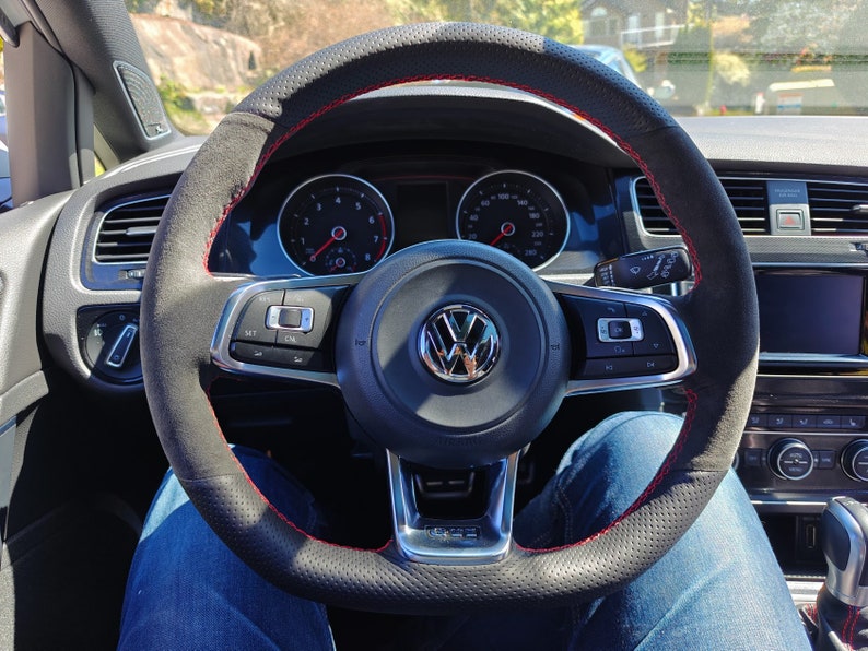 May include: A black leather steering wheel with red stitching and a Volkswagen logo in the center. The steering wheel has controls for the car's audio system and cruise control.