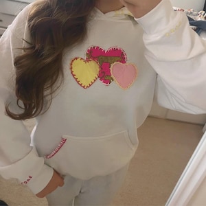 Op de afbeelding: Witte hoodie met een kangoeroezak en een capuchon met trekkoord. De voorkant heeft drie geborduurde harten in geel, roze en een luipaardprint. De manchet heeft geborduurde tekst. De zak en manchetten hebben rode stiksels.