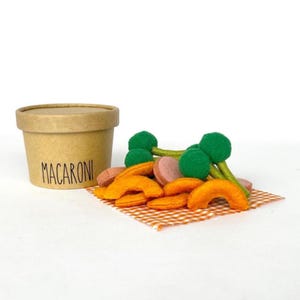 May include: A kraft paper cup labeled "MACARONI" sits next to felt food items. The felt food includes orange macaroni, pink sausage slices, and green broccoli florets, all arranged on a small orange and white checkered cloth.