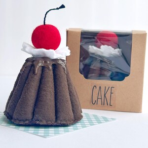 May include: A felt chocolate cake with white frosting and a red cherry on top. The cake is in a brown box with a clear window and the word "CAKE" printed on the front.