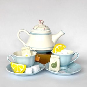 May include: A charming tea set featuring a white teapot with a floral lid, two light blue teacups with saucers, and a tea bag in each cup. The set includes lemon slices and sugar cubes, creating a delightful tea party scene.