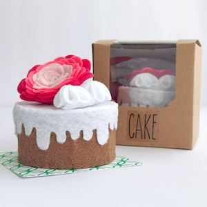 May include: A felt cake with a brown base, white frosting dripping down the sides, and a large pink and red flower on top. The cake sits on a green and white patterned paper. A boxed cake is visible in the background with the word "CAKE".