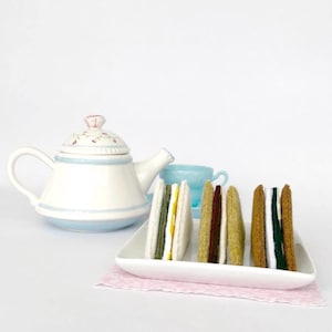 May include: A white teapot with a blue rim and floral accents sits next to a plate of felt sandwiches. A light blue teacup is in the background. The sandwiches have various fillings and are arranged on a white rectangular plate.