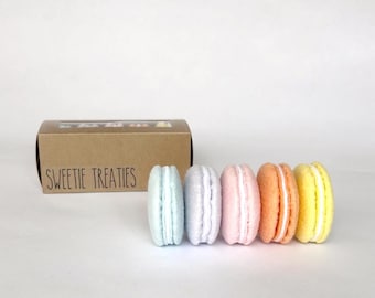 Puede incluir: Una caja de cartón marrón con una ventana transparente y el texto SWEETIE TREATIES impreso en el frente, acompañada de cinco objetos con forma de macarrón en tonos pastel azul claro, lavanda, rosa claro, naranja y amarillo con centros blancos.