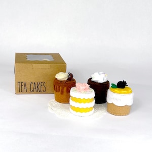 May include: A brown cardboard box with a window and the text TEA CAKES printed on the front, accompanied by four small felt cakes. The cakes feature various designs including a brown cake with white frosting, a yellow and white striped cake with a pink flower, a dark brown cake with white frosting, and a tan cake with white frosting and a yellow top.