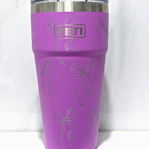 Op de afbeelding: Een paarse YETI Rambler tumbler met een deksel van roestvrij staal en een patroon van witte strikken. Het YETI-logo is afgedrukt op de voorkant van de tumbler.
