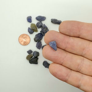 Puede incluir: Una colección de pequeñas piedras preciosas azul-grisáceas rugosas, algunas con un tono verdoso, esparcidas sobre una superficie blanca. Una sola piedra preciosa triangular más grande se sostiene en una mano para comparar el tamaño.