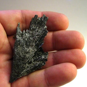 May include: A dark gray, naturally formed, mineral specimen with a pointed tip and a layered, fan-like structure.