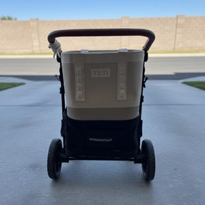 May include: A black stroller frame with a brown handle and a large beige YETI cooler bag attached to the back. The stroller features black wheels and a black storage compartment at the base with the white text WONDERFOLD printed on the front.