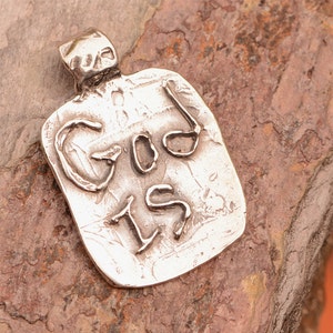May include: A silver pendant with the words "GOD IS" in a raised, textured font. The pendant is rectangular with a loop for a chain. It rests on a rough, reddish-brown stone. The pendant's design is simple and bold.