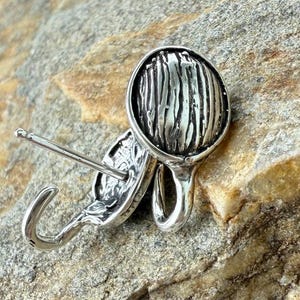 May include: A pair of silver-toned earrings. Each earring features a textured, circular design with vertical lines, connected to a curved hook and a post for piercing. The earrings are displayed on a textured, light brown stone surface.